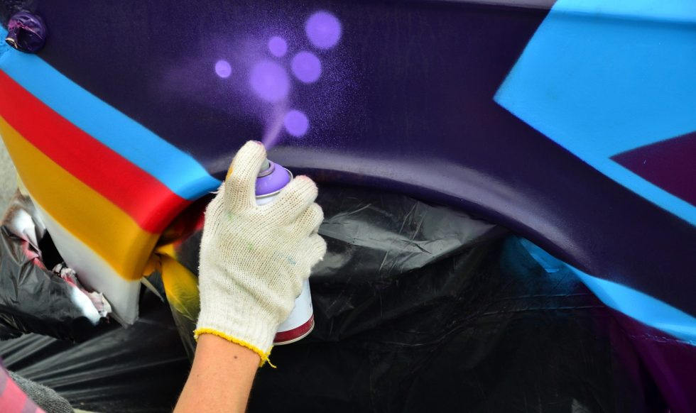 A hand with a spray can that draws a new colorful graffiti on the car. Photo of the process of drawing a graffiti on an automobile close-up. The concept of street art and illegal vandalism
